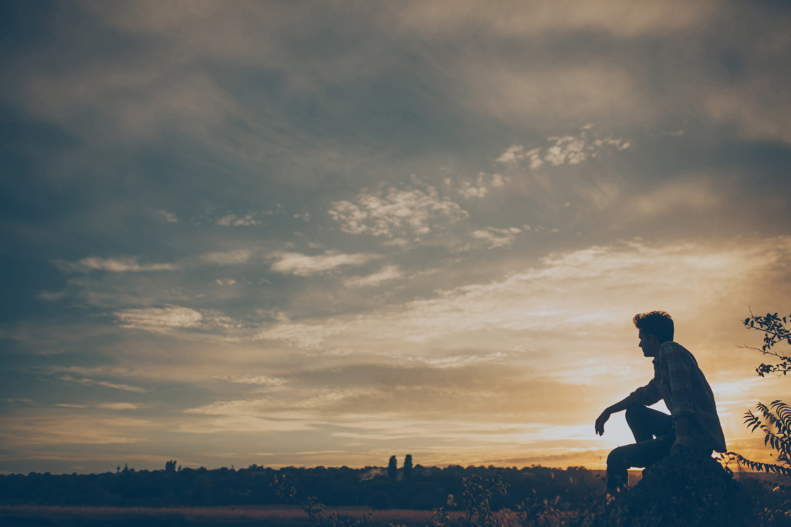 A partly cloudy sunset scene, with the silhouette of someone sitting in a field. They look to be deep in thought.