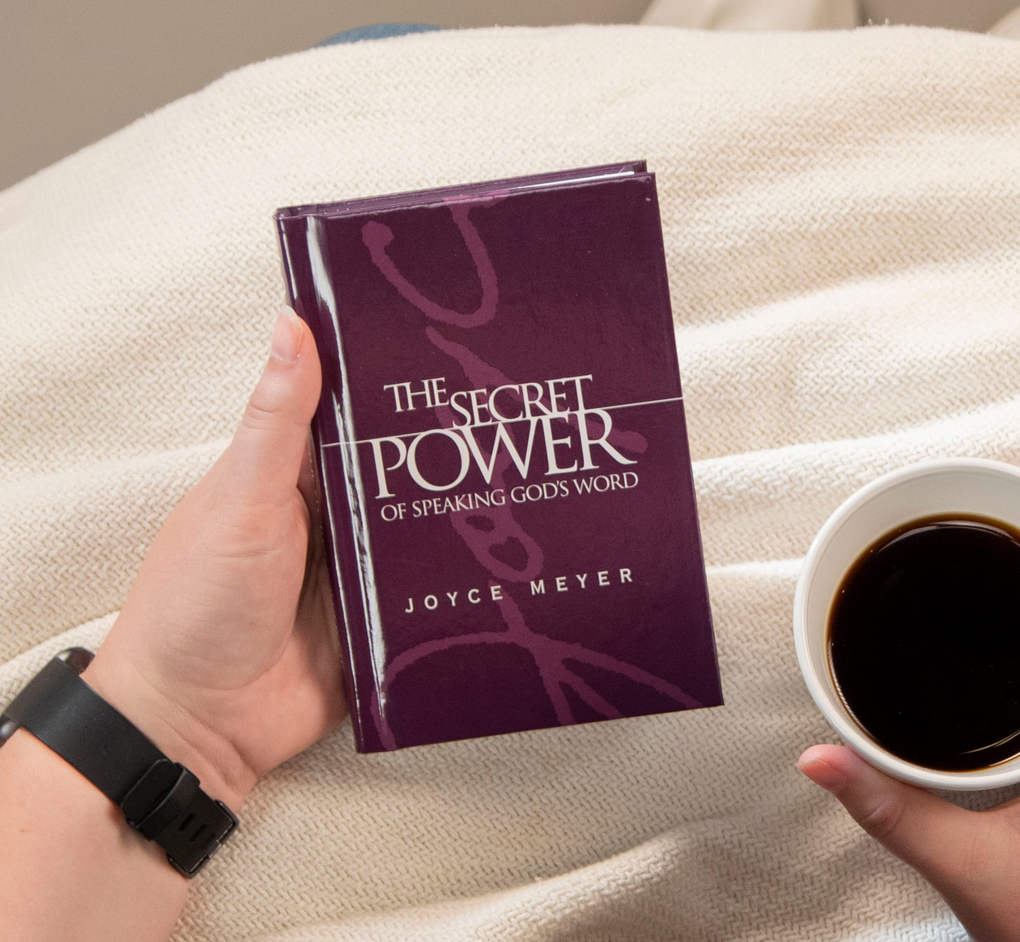 The Secret Power of Speaking God’s Word purple hardcover book being held in hand next to a cup of coffee